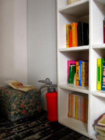 Corner of a modern dolls' house miniature pop-up Little Library, with a pouffe, fire extinguisher and shelves of books.
