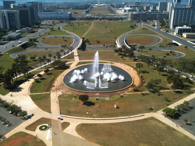 a fountain in a circular area with roads and buildings in the background