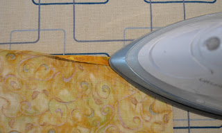 On the ironing board, the top hem has been folded once and the iron on the right hand side is pressing down the fold.
