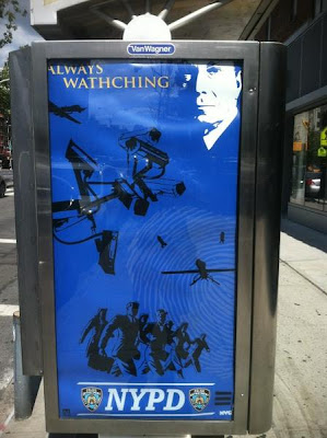 Blue poster in transit kiosk, with white image of Michael Bloomberg, cameras, and police in uniform with headline ALWAYS WATHCHING and the NYPD logo at bottom