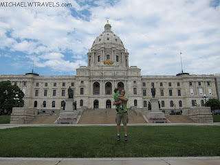 a man holding a child in front of a large white building with Minnesota State Capitol in the background