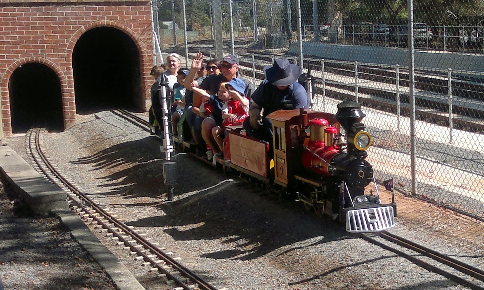 Model American style steam locomotive coming out of a tunnel. The loco is named "Yosemite Valley Railroad Jofesine No. 9"  travelling with two carriages loaded with passengers.  The full-scale 'real-life' suburban railway lines can be seen beyond the fence.