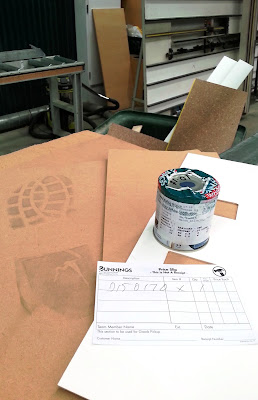 Several pieces of MDF (one with a large boot print on it) and a test pot of paint in front of a bin in a hardware store.