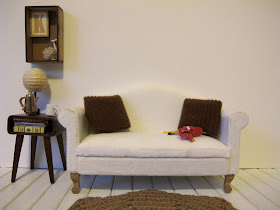 Modern dolls' house miniature lounge scene of a sofa and side table. On the sofa is some knitting and on the side table is a  lamp, coffee pot and mug. On the wall above it is a box frame with the letter K, a ball of wool and two knitting needles.