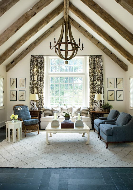 Living room in a pool house with high beamed ceiling, wine barrel chandelier, slate floors, diamond patterned white rug, blue armchairs, a white sofas, graphic print curtains, a white coffee table and one brown armchair