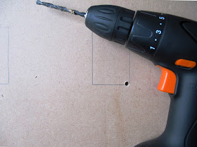 Electric drill on a piece of MDF board, with two rectangles drawn on it, one of which has a hole drilled in the corner of it.