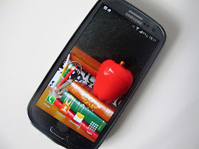 Mobile phone withg wallpaper depicting a modern dolls' house scene of a pile of books with a large apple on top, and a jar of pens and pencils next to it.