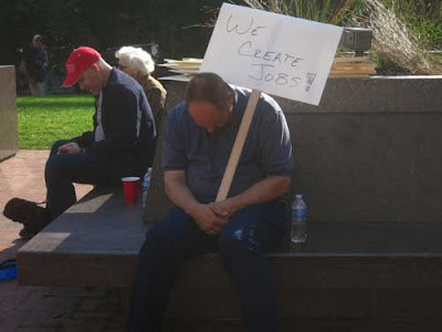 Man sitting, slumped and possibly asleep, holding a sign on his shoulder that says We Create Jobs