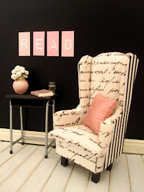 Modern doll's house miniature scene of a black and white wing chair against a black wall. On the wall are the letters R,E,A and D in pink and white and on the table under them is a pink vase, book and a glass of water.
