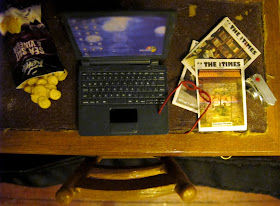 Modern dolls' house miniature desk, from above, showing an opened bag of chips, a laptop, several issues of The tiny Times magazine, red reading glasses and a digital camera.