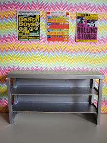 Selection of bright 1960s band posters arranged on a bright, zig zag-patterned wall. Underneath is a modern doll's house miniature industrial grey storage bench.