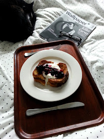 Cat curled up asleep on a bed next to a magazine and a tray holding a plate of french toast, yoghurt and berries.