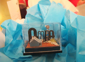 A small display box containing the letters 'AMEA', and a tiny cottage and windmill, displayed on some scrunched-up tissue paper.