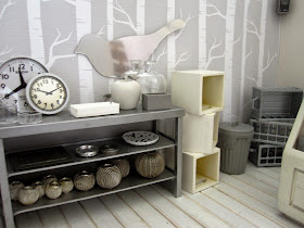 View of a modern dolls' house miniature homeware shop in grey and white. On display is  a grey metal shelving unit with various grey-coloured decor pieces. To the right is a pile of white storage boxes, a grey trash can and a pile of grey storage baskets.