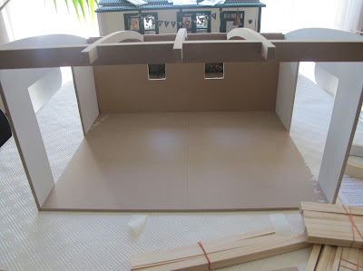 A half-built dolls' house shed from the front, with a collection of wooden lengths in front.