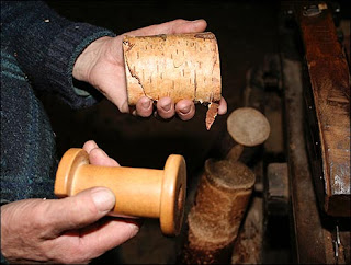 Close up of a man's hands holding a short section of silver birch wood (with bark) in one hand and a finished bobbin in the other.
