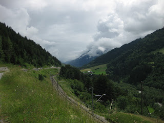 Oberalpstrasse snakes through the valley above the railroad tracks, with dramatic clouds sweeping up from a distant peak, Switzerland