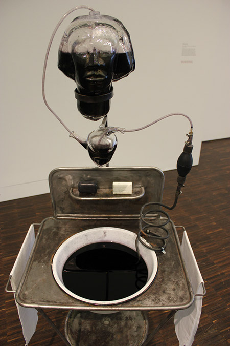 Alison Saar - Still... Exhibit at the Figge Museum, Davenport, Iowa. Photograph by Robin Peters of a Sink filled with a black liquid. Above the sink, suspended by metal "still-like" tubing is a glass heart and above that a glass head half filled with black liquid. A bar of white soap and black soap on the old styled sink.