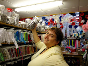 Woman in a haberdashery section of a craft shop, holding up a large jar of studs.