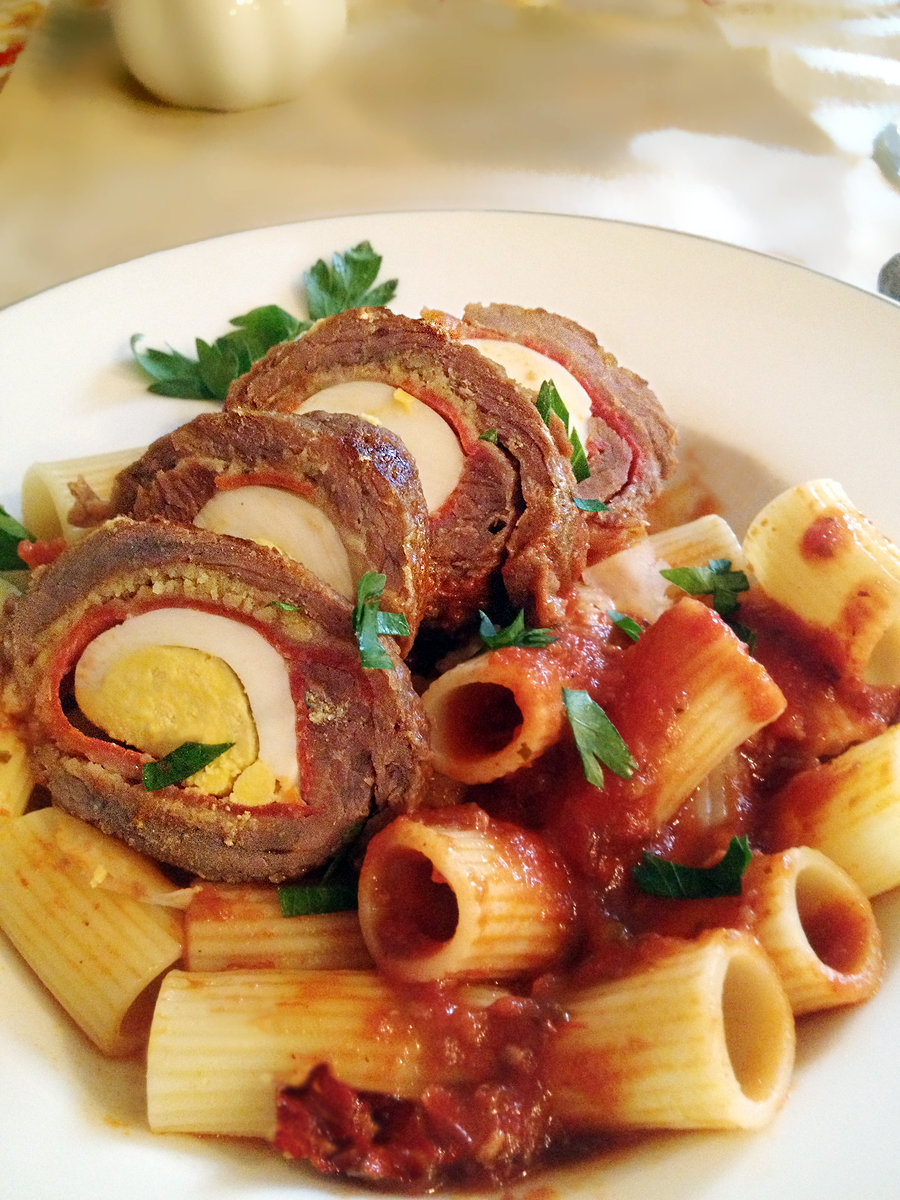 Braciole sliced to show the egg inside, atop a serving of rigatoni with tomato sauce.
