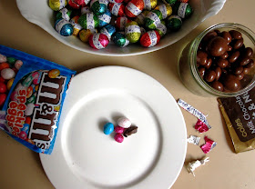 A bag of M&M speckled eggs, a bowl of Cadbury mini eggs and a jar of milk chocolate fruit and nut mix arranged around a plate with dolls' house-sized Easter eggs on it.