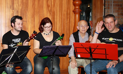Four people, holding ukuleles. I am looking down and smiling - someone else is grinning and waving at the camera.