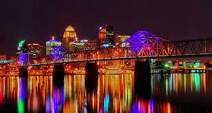 colorful photo of the big four bridge lit up at night with blue, green ornge lights reflecting into the river and the large buildings in the background