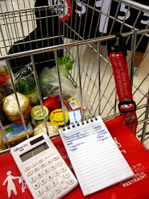 Supermarket trolley with groceries in it, a calculator and grociery list on the child seat. Behind the trolley is a sign which says '1/2 price: $1.55)