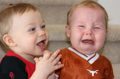 a toddler wearing an OU shirt clapping his hands while another toddler wearing a Texas shirt is crying.