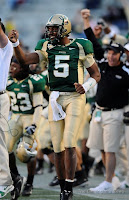 A football player wearing gold pants, green jersey and a dragon his gold helmet celebrates on the sidelines of a football game.