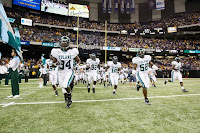 Football players in white Tulane uniforms take an artificial turf football field before a game with fans in the stands.
