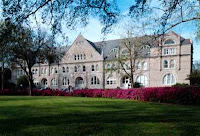 Large three story pink brick building with rose bushes an expansive green lawn and trees in front of the academic building.