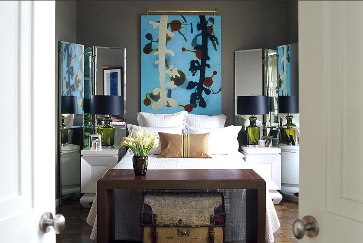 Dark grey bedroom with wood floor, white side tables with mirrored screens behind them, a piece of blue modern art instead of a headboard and a wood table at the foot of the bed with a trunk underneath