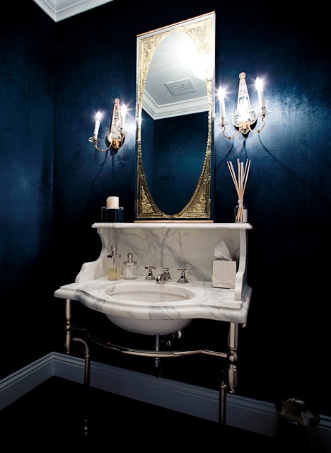 Bathroom with navy blue walls, a large art deco gilded mirror, two wall mounted candlesticks, dark wood floor, moulded ceiling and a marble console sink