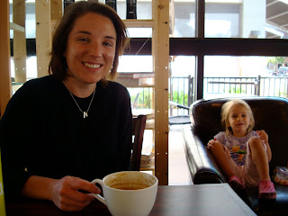Woman sitting at table with large mug of coffee and young girl sitting in chair in background