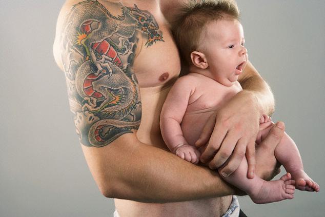 A man holding his baby has a beautiful dragon tattoo on his arm to shoulder.