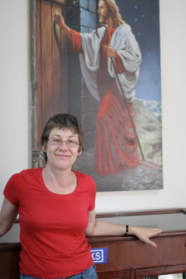 Woman with short hair and red t-shirt standing in front of a painting of Jesus