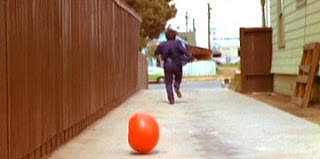 Em um pátio ao lado de uma casa, um homem corre fugindo de um enorme tomate que rola atrás dele. O homem está de costas, correndo para longe e o tomate está em primeiro plano.