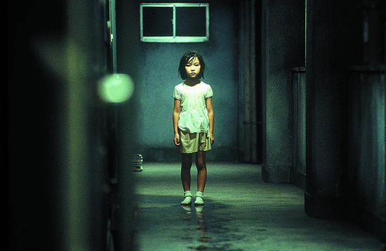 Uma menininha japonesa está parada quase no fim de um corredor. O Chão e paredes são acinzentados e com aspecto sujo, a luz é fria. A garotinha está séria, usa camiseta branca, short, meias brancas e seus cabelos curtos estão molhados. O chão está molhado perto dela.