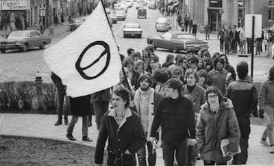 Black and white photo of a small group of young people marching up some stairs, carrying a white flag with the black ecology symbol on it