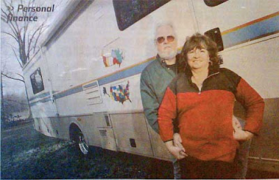 Man and woman in front of their RV. The woman is wearing a sweater that is mostly dark red with black shoulders