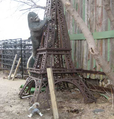 A large plush King Kong climbs a rusted steel Eiffel Tower, while a small Rambo figure attempts to climb up the bottom leg of the tower