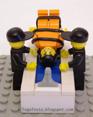 Lego figures of two guys in black clothes and hats tipping another guy (strapped to a table, wearing an orange jumpsuit) into some water