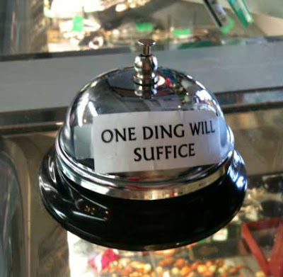 Metal service bell on a counter with a small sign taped on reading ONE DING WILL SUFFICE