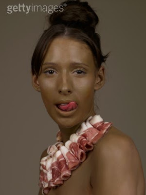 Young white woman with skin painted brown, wearing a bacon necklace and licking her lips