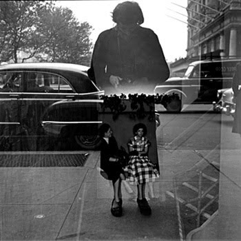 Reflected window photo where Maier is seen in the glass with her camera, two a man and woman, seated, appear to be inside the lower part of her coat