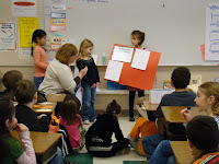 A young student holding an orange poster with pages of her writing on it. Another young student speaking into the microphone the teacher is holding while the class sits and listens to their presentation