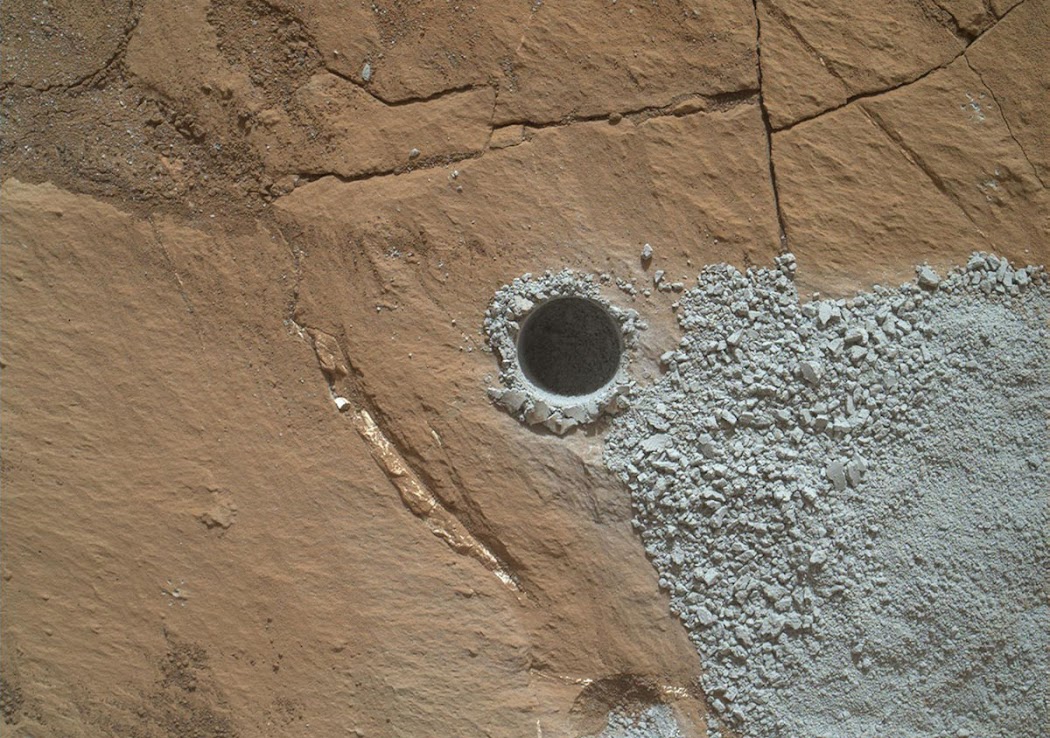 Một lỗ được tạo ra sau khi tàu Curiosity thực hiện khoan xuống để thu thập vật chất từ một phần của tảng đá lớn được gọi là Buckskin vào ngày 30 tháng 7 năm 2015, hay sol 1060. Đường kính của lỗ này nhỏ hơn đồng xu một chút. Bột đá được lấy ra sau khi khoan được đưa vào thiết bị phân tích tích hợp trong tàu. Việc khoan lỗ ở tảng đá Buckskin không được lên kế hoạch từ trước, tàu cũng không có kinh nghiệm để khoan lỗ ở nơi này. Vào đầu năm 2015, Curiosity phát hiện mức độ silic và hydro cao bất thường ở đây, một khu vực gần Đèo Marias ở chân núi Sharp, nên các nhà khoa học mặt đất đã thay đổi một chút trong kế hoạch để biết được chuyện gì đang xảy ra. Hình ảnh: JPL-Caltech/MSSS/NASA. Một lỗ được tạo ra sau khi tàu Curiosity thực hiện khoan xuống để thu thập vật chất từ một phần của tảng đá lớn được gọi là Buckskin vào ngày 30 tháng 7 năm 2015, hay sol 1060. Đường kính của lỗ này nhỏ hơn đồng xu một chút. Bột đá được lấy ra sau khi khoan được đưa vào thiết bị phân tích tích hợp trong tàu. Việc khoan lỗ ở tảng đá Buckskin không được lên kế hoạch từ trước, tàu cũng không có kinh nghiệm để khoan lỗ ở nơi này. Vào đầu năm 2015, Curiosity phát hiện mức độ silic và hydro cao bất thường ở đây, một khu vực gần Đèo Marias ở chân núi Sharp, nên các nhà khoa học mặt đất đã thay đổi một chút trong kế hoạch để biết được chuyện gì đang xảy ra. Hình ảnh: JPL-Caltech/MSSS/NASA.