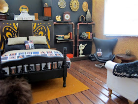 One-twelfth scale miniature scene, with a bed dressed in linen and black with throw rug and cushion printed with letter As, in front of a false wall. To the right is a lounge area with a set of pipe shelves, a black egg chair and a cream sofa.