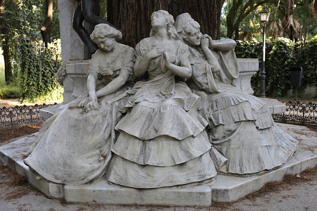 Vista de tres estatuas femeninas completas de piedra blanca sentadas en un árbol.